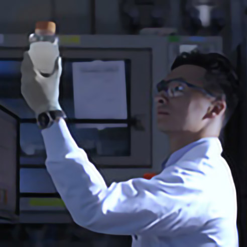 Ein Forscher mit Schutzbrille und Handschuh hält ein Probengefäß im Labor gegen das Licht zur Untersuchung. Symbolbild für Kontaktdaten des Kundenservices.