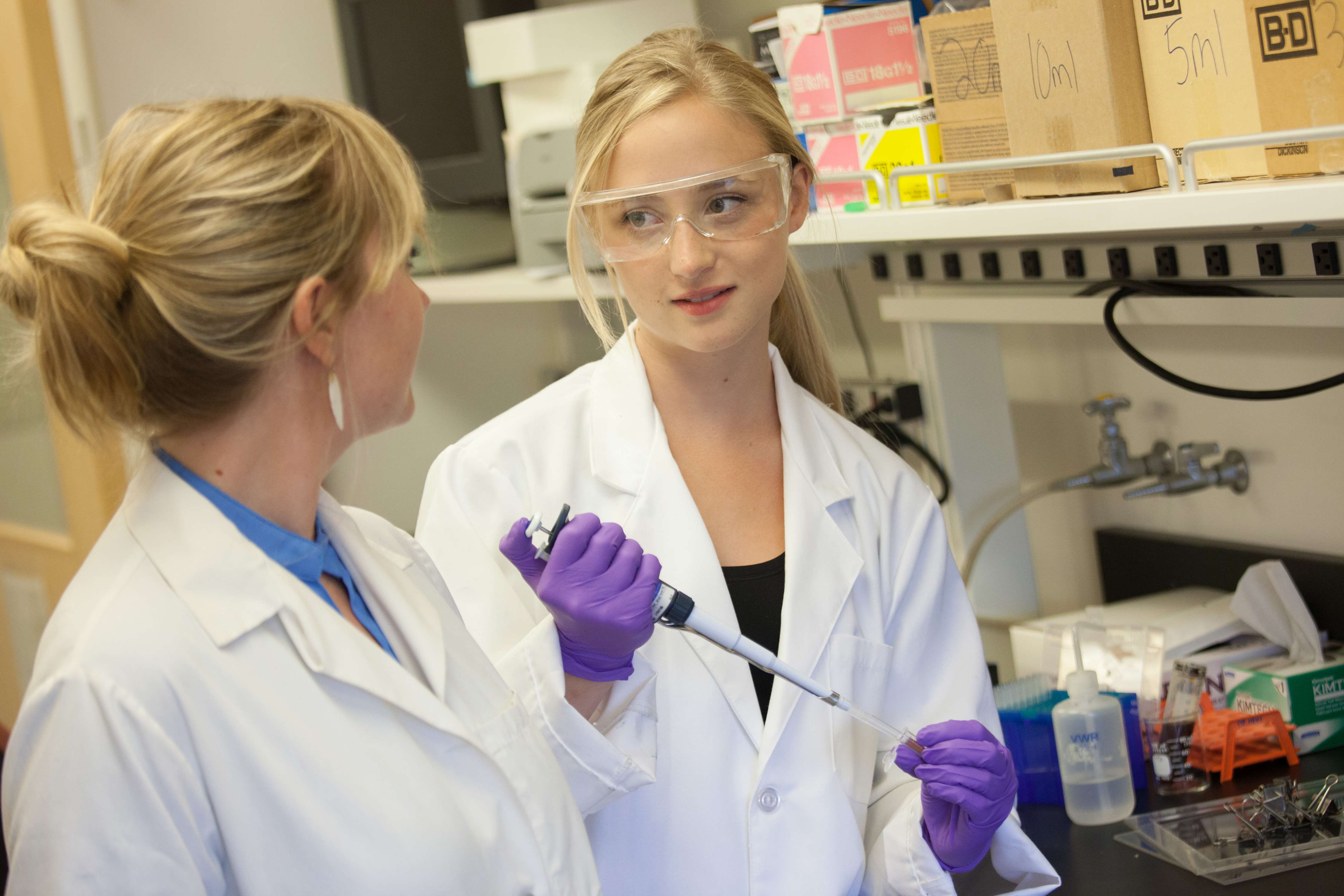 Zwei junge Frauen pipettieren im Labor eine Lösung und unterhalten sich. Sie tragen weiße Kittel und lila Gummihandschuhe.