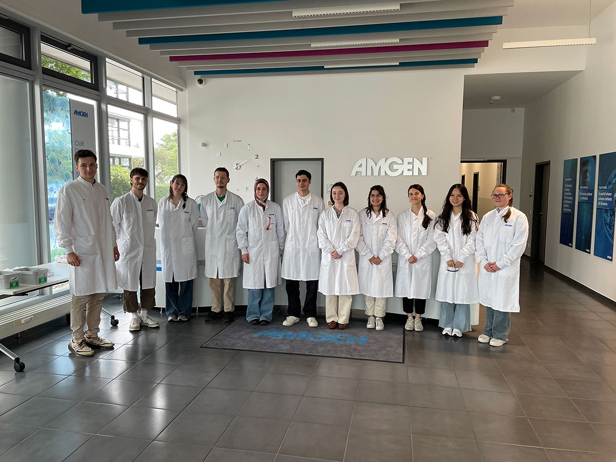 Elf junge Studierende in wei&szlig;en Kitteln stehen aufgereiht in einem Eingansbereich und l&auml;cheln in die Kamera. Im Hintergrund sind das Amgen Logo und einige Plakate zu sehen.