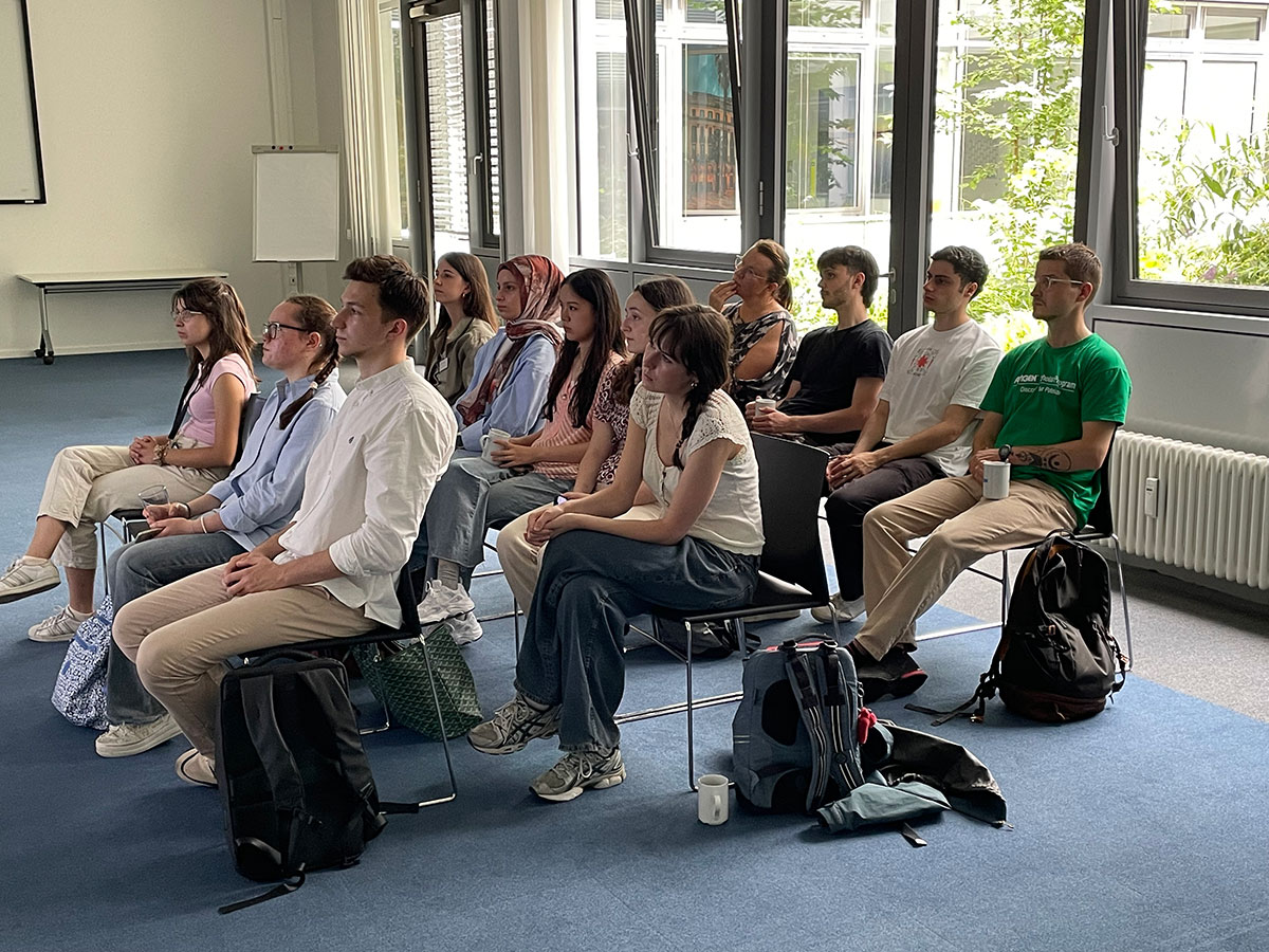 Eine Gruppe von zw&ouml;lf jungen Studierenden sitzt in drei Reihen auf St&uuml;hlen in einem hellen Raum mit gro&szlig;en Fenstern. Die Personen sind aufmerksam nach vorne gerichtet, als w&uuml;rden sie einer Pr&auml;sentation oder einem Vortrag folgen.