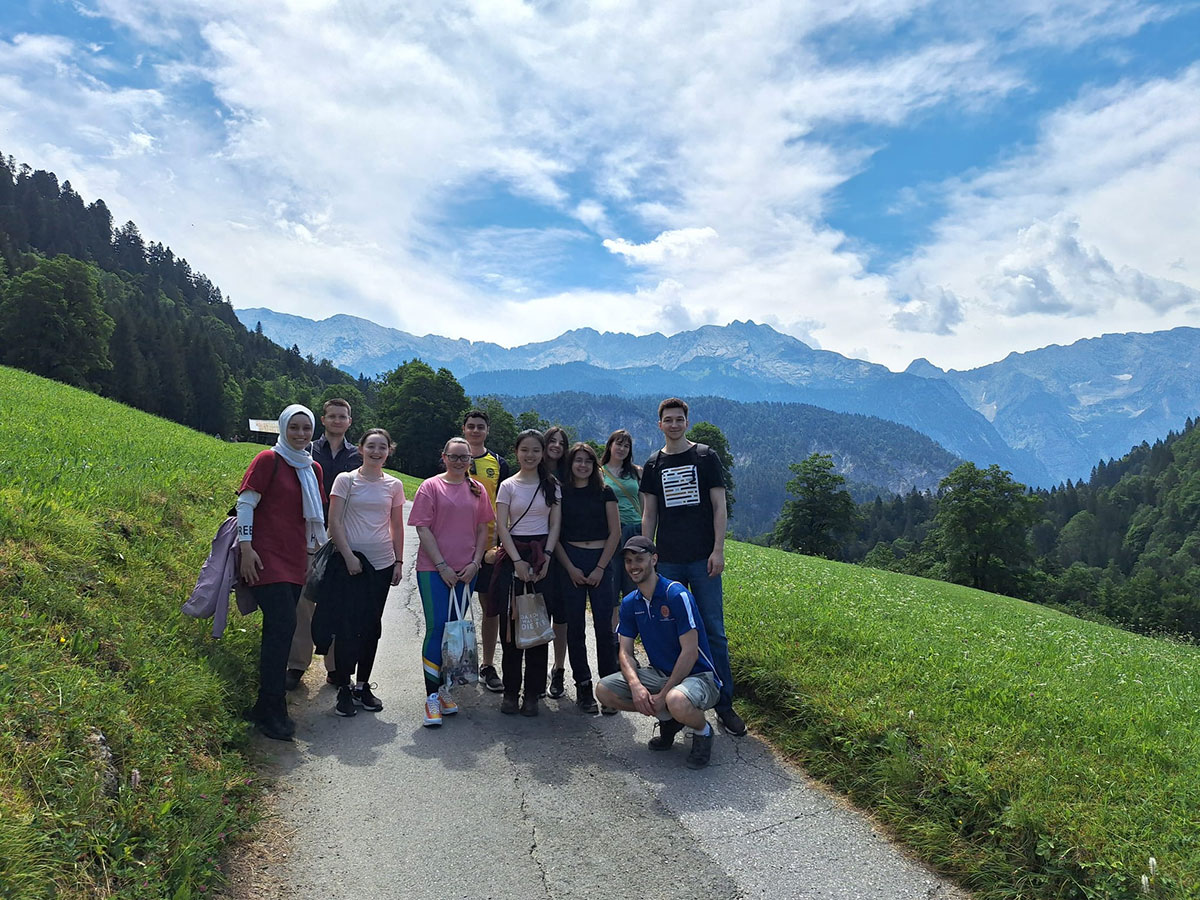 Eine Gruppe von elf Studierenden steht auf einem schmalen Weg in einer gr&uuml;nen Berglandschaft mit Wiesen, W&auml;ldern und hohen Bergen im Hintergrund unter blauem Himmel mit Wolken.